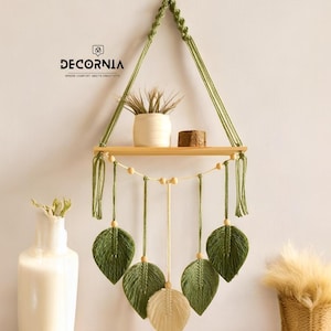 Puede incluir: Estante de pared de macramé con un estante de madera y hojas verdes colgantes. El estante está suspendido por una cuerda verde y presenta una pequeña planta en una maceta blanca y un pequeño bloque de madera. Para la decoración del hogar.