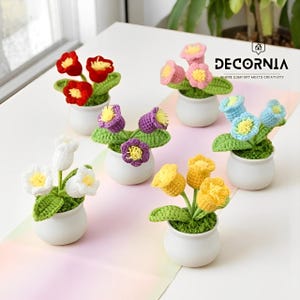 Könnte beinhalten: Sechs kleine, weiße Keramiktöpfe, jeweils mit einem gehäkelten Blumenarrangement. Die Blumen sind in verschiedenen Farben gehalten: rot, rosa, lila, blau, gelb und weiß, mit grünen Blättern. Das Logo "DECORNIA" ist sichtbar.