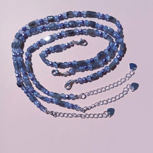 Può includere: Set di collane a strati con più fili di perline di pietre preziose blu e prolunghe a catena argentate. Le perline variano nella forma, tra cui rettangolare e rotonda, con una miscela di tonalità blu chiare e scure. La collana è esposta su uno sfondo rosa.