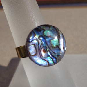 Pode incluir: Anel de concha de abalone com um cabochão redondo iridescente azul, verde e branco. O anel tem uma banda dourada. A concha de abalone tem um padrão de redemoinho. O anel está em uma superfície branca.