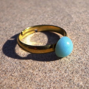 Pode incluir: Um anel ajustável dourado com uma pedra azul clara em forma de ovo. O anel está sobre uma superfície texturizada castanho-clara, com a sombra do sol.