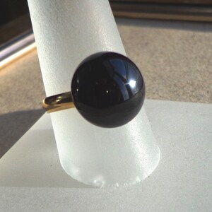 Pode incluir: Um anel dourado com uma grande esfera preta brilhante como peça central. A esfera reflete a luz, criando destaques. O anel é exibido em um suporte branco em forma de cone, destacando o design da joia.