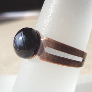 Op de afbeelding: Een koperkleurige ring met een grote, ronde, glanzende zwarte steen. De ringband heeft een uniek open ontwerp. De ring wordt getoond op een wit, cilindrisch object.