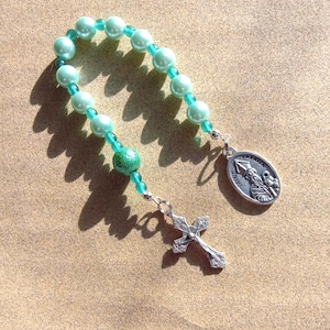 May include: A single-decade rosary featuring light mint green pearl-like beads separated by smaller translucent green beads. A textured green bead sits near the bottom, connecting to a silver-toned crucifix and an oval medal depicting Saint Patrick.