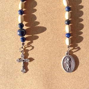 May include: A rosary with alternating light-colored and dark blue beads. The rosary features a silver-colored crucifix and a pendant with a religious figure. The beads are strung together with a silver-colored chain.