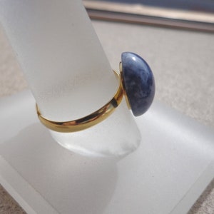 Pode incluir: Um anel dourado com uma grande pedra preciosa azul em forma de meia cúpula. A pedra preciosa tem uma aparência marmorizada com tons de azul e branco. O anel é exibido em um suporte transparente e inclinado.