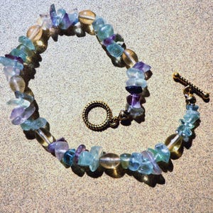 May include: A bracelet composed of irregular, tumbled gemstone chips in shades of translucent light blue, purple, and pale yellow. The piece features a gold-toned metal toggle clasp with a textured, twisted rope design on both the ring and bar.