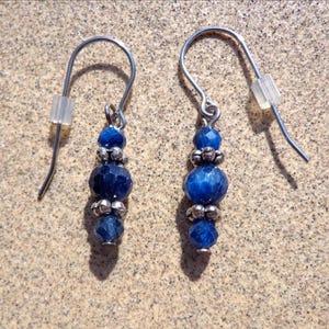 May include: A pair of drop earrings featuring faceted deep blue beads arranged in a vertical line, separated by small silver-toned metal spacers. Each earring hangs from a curved silver-toned wire hook and includes a clear plastic backing.
