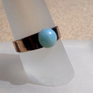Pode incluir: Um anel em tom de cobre com uma pedra redonda azul clara. O anel é exibido em um suporte branco inclinado. A pedra está centralizada na banda, criando um design simples e elegante. A banda do anel tem uma aparência escura e texturizada.