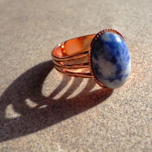 Könnte beinhalten: Kupferring mit einem großen, ovalen blauen Sodalith-Stein. Der Stein hat weiße Marmorierungen. Der Ring hat ein dreifaches Banddesign und eine strukturierte Lünettenfassung. Der Ring wird auf einer neutralen Oberfläche mit einem ausgeprägten Schatten dargestellt.
