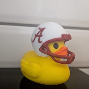 Puede incluir: Un pato de goma amarillo con un casco de fútbol blanco con un logotipo "A" carmesí y una máscara facial a juego. El pato tiene un pico naranja y ojos negros. Un artículo divertido y novedoso.
