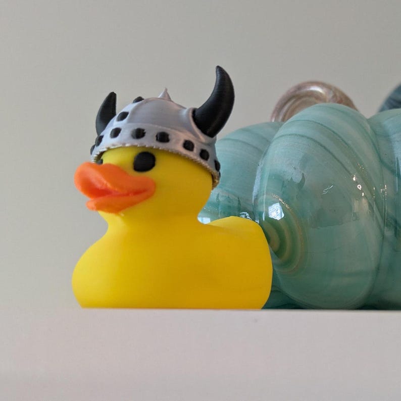 May include: A yellow rubber duck wearing a silver Viking helmet with black horns. The duck has an orange beak and black eyes. A teal glass pumpkin is in the background.