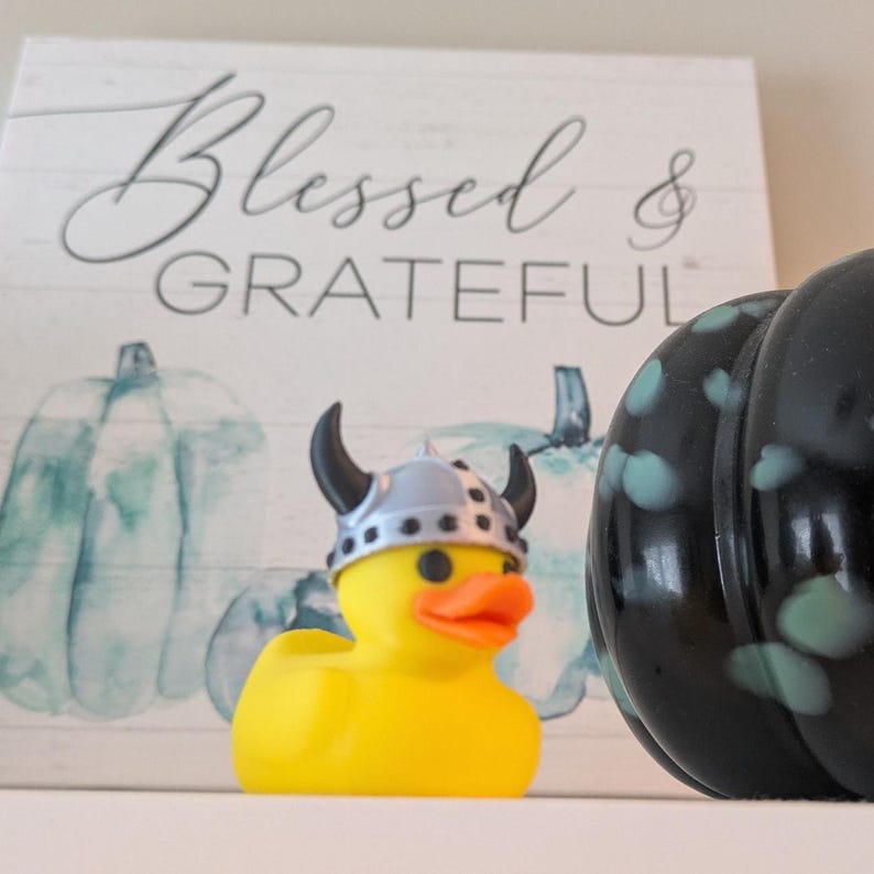 May include: A yellow rubber duck wearing a silver Viking helmet with black horns and an orange beak. The duck is in front of a sign that says "Blessed & Grateful" and a black pumpkin with teal spots.