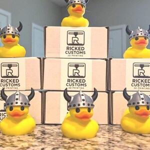 May include: Yellow rubber duck toys wearing silver Viking helmets with black horns. The ducks are arranged in front of boxes with the text "RICKED CUSTOMS 3D PRINTING" printed on them.
