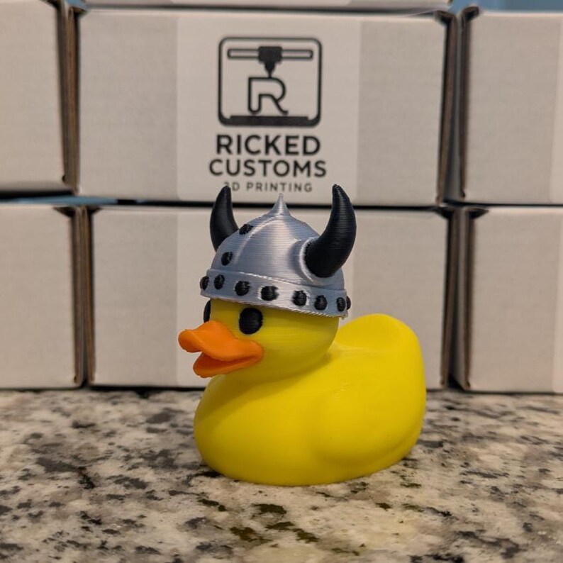 May include: A yellow rubber duck wearing a silver Viking helmet with black horns. The duck has an orange beak and black eyes. The background includes boxes with the text "RICKED CUSTOMS 3D PRINTING".