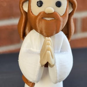 May include: A 3D-printed figurine of Jesus with brown hair and beard, wearing a white robe. His hands are clasped in prayer. The figurine has a cartoonish design and is set against a blurred brick background.