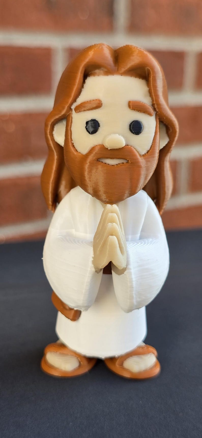 May include: A 3D-printed figurine of Jesus with brown hair and beard, wearing a white robe. His hands are clasped in prayer. The figurine has a cartoonish design and is set against a blurred brick background.
