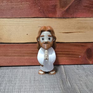 May include: A small, stylized figurine of Jesus with brown hair and a beard, wearing a white robe. The figure has its hands clasped in prayer and is standing on a wooden surface. The figurine is approximately 4 inches tall.