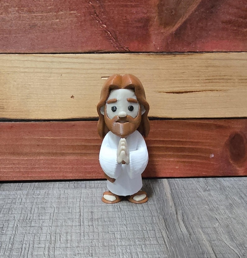 May include: A small, stylized figurine of Jesus with brown hair and a beard, wearing a white robe. The figure has its hands clasped in prayer and is standing on a wooden surface. The figurine is approximately 4 inches tall.