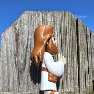 May include: A 3D-printed figurine of a religious figure, depicted in a white robe with brown hair and beard, and sandals. The figure is posed with hands together in prayer. The background features a weathered wooden fence and a clear blue sky.