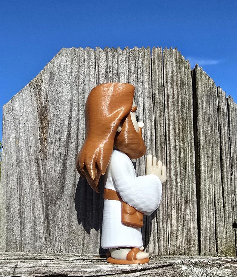 May include: A 3D-printed figurine of a religious figure, depicted in a white robe with brown hair and beard, and sandals. The figure is posed with hands together in prayer. The background features a weathered wooden fence and a clear blue sky.