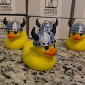 May include: A group of yellow rubber ducks wearing silver Viking helmets with black horns. The ducks have orange beaks and black eyes. The background features boxes with the text "Ricked Customs 3D Printing."