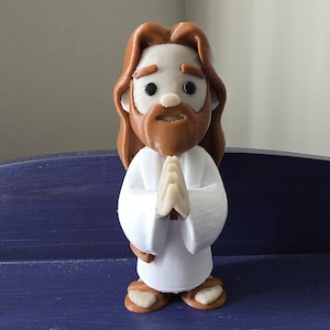 May include: A figurine of Jesus with brown hair and beard, wearing a white robe. His hands are clasped in prayer. The figure has a light skin tone and is standing on a blue surface.