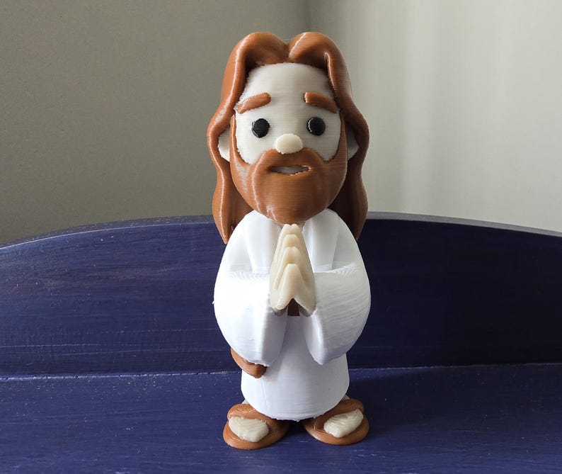 May include: A figurine of Jesus with brown hair and beard, wearing a white robe. His hands are clasped in prayer. The figure has a light skin tone and is standing on a blue surface.