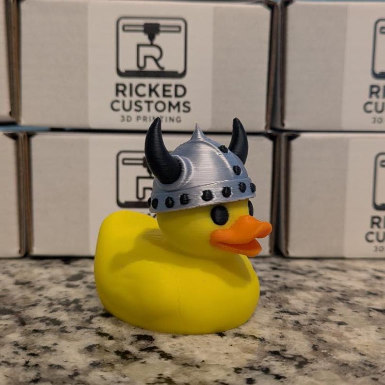 May include: A yellow rubber duck wearing a silver and black Viking helmet. The duck has an orange beak and black eyes. The background features boxes with the text "RICKED CUSTOMS 3D PRINTING".