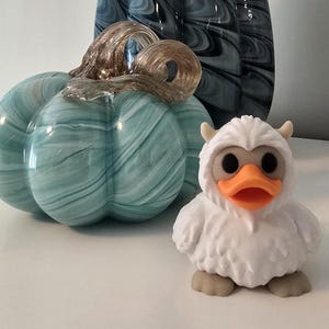 May include: A teal glass pumpkin with a gold stem sits next to a white Yeti duck figurine. The pumpkin has a swirled pattern. The duck has an orange beak, black eyes, and small horns.