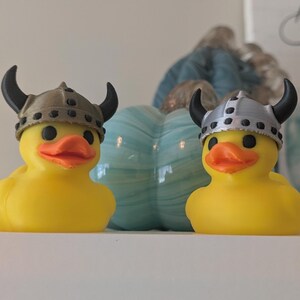 May include: Two yellow rubber ducks wearing Viking helmets with black horns. The ducks have orange beaks and black eyes. A light blue pumpkin-shaped decoration is behind them.
