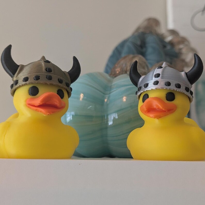 May include: Two yellow rubber ducks wearing Viking helmets with black horns. The ducks have orange beaks and black eyes. A light blue pumpkin-shaped decoration is behind them.