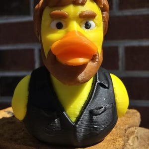 Op de afbeelding: Een gele badeend met bruin haar en baard, gekleed in een zwart vest. De eend heeft een oranje snavel en ligt op een steen. De achtergrond is een bakstenen muur.
