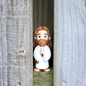 May include: A figurine of Jesus with brown hair and a beard, wearing a white robe, and with hands clasped in prayer. The figurine is standing between two weathered wooden panels.