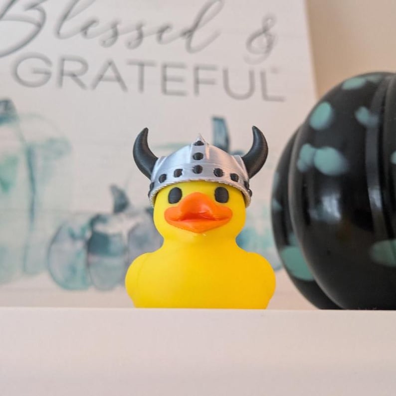 May include: A yellow rubber duck wearing a silver Viking helmet with black horns and an orange beak. The duck is in front of a blurred background with the words "Blessed & Grateful".