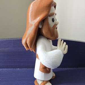 May include: A 3D-printed figurine of a religious figure with long brown hair and a beard, wearing a white robe and brown sandals. The figure has its hands raised in a gesture of prayer or blessing. The figurine is a light brown and white color.