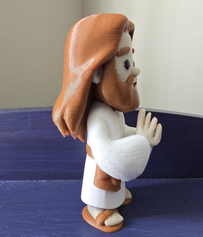May include: A 3D-printed figurine of a religious figure with long brown hair and a beard, wearing a white robe and brown sandals. The figure has its hands raised in a gesture of prayer or blessing. The figurine is a light brown and white color.