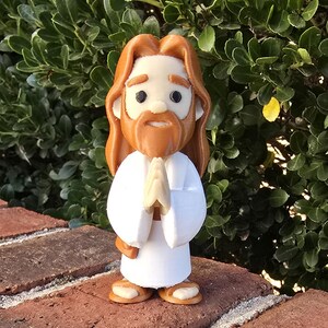 May include: A figurine of a religious figure with long brown hair and a beard, wearing a white robe. The figure has hands clasped in prayer and is standing on a brick surface. The background is a green bush.
