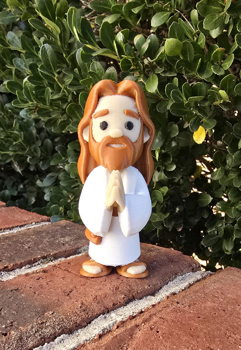 May include: A figurine of a religious figure with long brown hair and a beard, wearing a white robe. The figure has hands clasped in prayer and is standing on a brick surface. The background is a green bush.