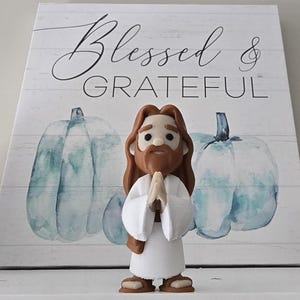 May include: A figurine of Jesus with brown hair and a beard, wearing a white robe, with hands clasped in prayer. The figurine is in front of a wooden sign with the words "Blessed & GRATEFUL" and two blue watercolor pumpkins.