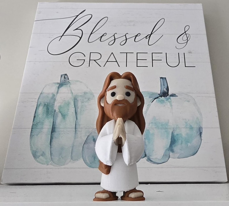 May include: A figurine of Jesus with brown hair and a beard, wearing a white robe, with hands clasped in prayer. The figurine is in front of a wooden sign with the words "Blessed & GRATEFUL" and two blue watercolor pumpkins.