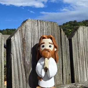May include: A 3D-printed figurine of Jesus, depicted with long brown hair and a beard, wearing a white robe. The figure has its hands clasped in prayer. The figurine is set against a weathered wooden fence with a blue sky and clouds in the background.