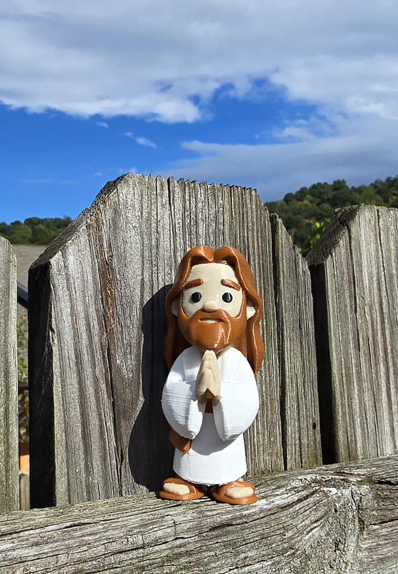 May include: A 3D-printed figurine of Jesus, depicted with long brown hair and a beard, wearing a white robe. The figure has its hands clasped in prayer. The figurine is set against a weathered wooden fence with a blue sky and clouds in the background.