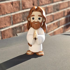 May include: A small, stylized figurine of Jesus with brown hair and a beard, wearing a white robe. His hands are clasped in prayer. The figure has a cartoonish design and is standing on a gray surface.
