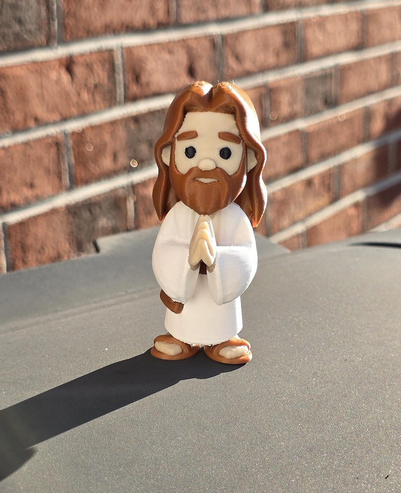 May include: A small, stylized figurine of Jesus with brown hair and a beard, wearing a white robe. His hands are clasped in prayer. The figure has a cartoonish design and is standing on a gray surface.