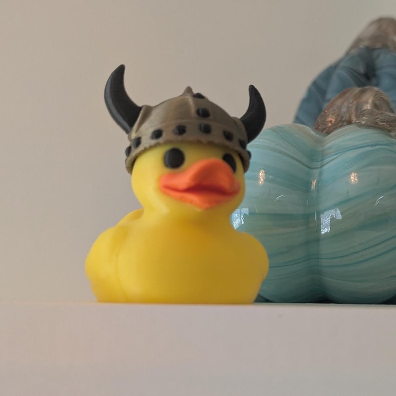 May include: A yellow rubber duck wearing a Viking helmet with black horns. The duck has black eyes and an orange beak. A blue and white decorative pumpkin is in the background.