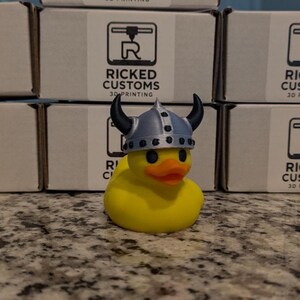 May include: A yellow rubber duck wearing a silver and black Viking helmet. The duck has an orange beak and black eyes. The background includes boxes with the text "RICKED CUSTOMS 3D PRINTING".