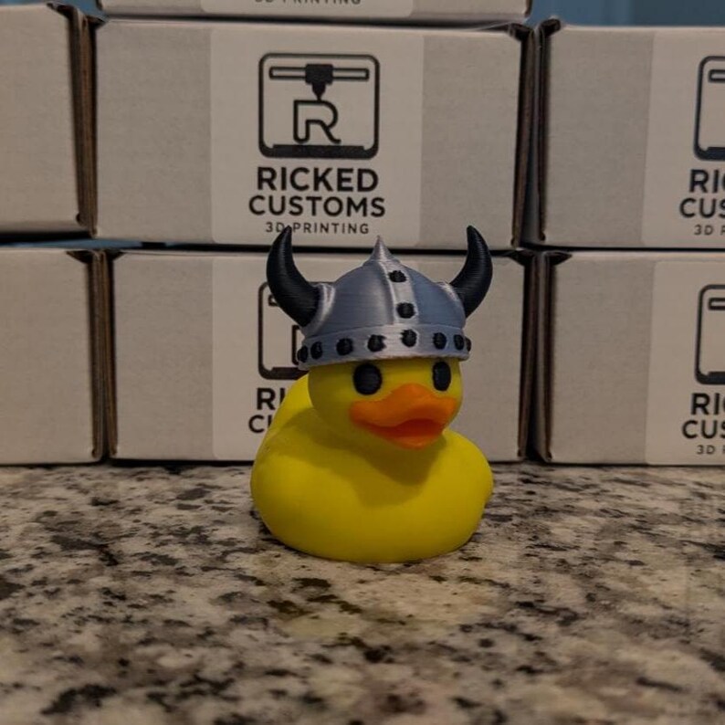 May include: A yellow rubber duck wearing a silver and black Viking helmet. The duck has an orange beak and black eyes. The background includes boxes with the text "RICKED CUSTOMS 3D PRINTING".