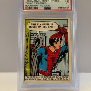 May include: A 1966 Marvel Super Heroes Spider-Man trading card, graded and encased. The card features Spider-Man in a red and blue costume, with the text "THIS FLY PAPER IS ROUGH ON THE HAIR!" in a speech bubble. The card is in a plastic holder.