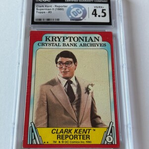 May include: A graded trading card featuring Clark Kent from Superman II (1980). The card has a red border and a photo of Clark Kent in a suit. The card is graded 4.5 by CGC. Text includes "KRYPTONIAN CRYSTAL BANK ARCHIVES" and "CLARK KENT REPORTER".